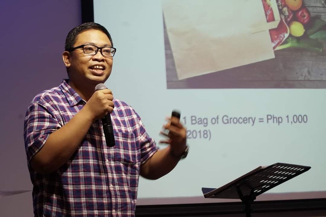 Kuya Aries speaking at a financial literacy talk, explaining the real cost of a bag of groceries